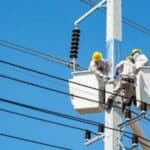 Electricians wearing safety gear work on power lines using lifts against a clear blue sky.